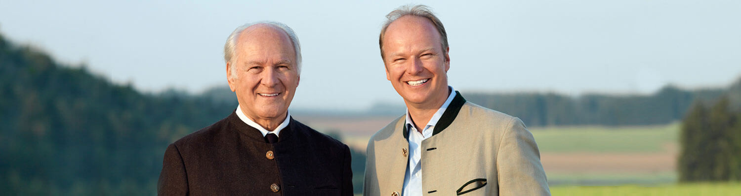 Two men, one older with white hair and the other younger with brown hair, stand side by side outdoors.